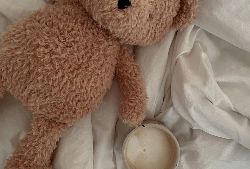 A well-loved teddy bear lying on crumpled white bedding next to a warm mug, suggesting comfort and retreat after a difficult day